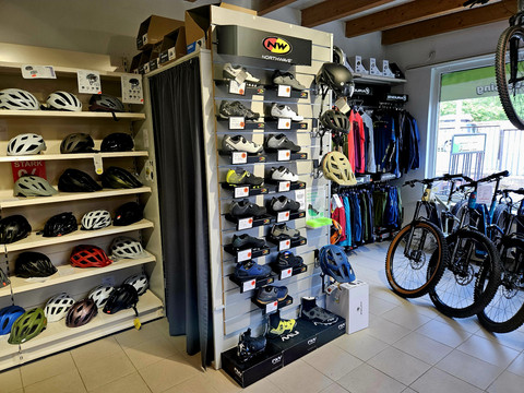 Verkoopruimte met verschillende modellen Fahrradgeschäft mit Regalen voller Helme und Schuhe, daneben hängen bunte Jacken; rechts stehen mehrere Fahrräder.