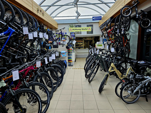 Fahrradgeschäft mit zahlreichen Fahrrädern in verschiedenen Farben und Größen, aufgereiht entlang der Wände; Schild "Achtung Stufe!" im Hintergrund.