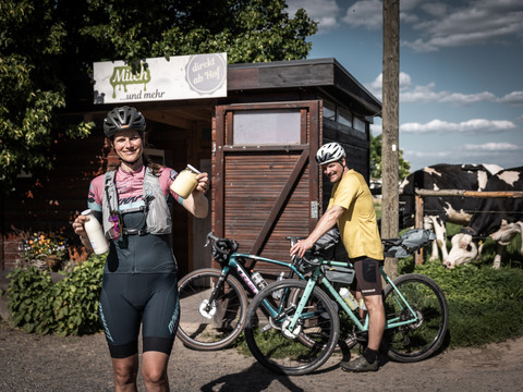 Fresh local products from Milchhof Fiedler Frau in Radkleidung hält Milchflaschen vor einem Hofladen, daneben ein Mann mit Fahrrad; im Hintergrund Kühe auf der Weide.Woman in cycling clothes holding milk bottles in front of a farm store, next to her a man with a bicycle; in the background cows in the pasture.Žena v cyklistickém oblečení s lahvemi mléka před farmářským obchodem, vedle ní muž s kolem; v pozadí krávy na pastvině.Kobieta w stroju kolarskim trzymająca butelki z mlekiem przed sklepem rolniczym, obok niej mężczyzna na rowerze; w tle krowy na pastwisku.Vrouw in fietskleding met melkflessen voor een boerderijwinkel, naast haar een man met fiets; op de achtergrond koeien in de wei.Donna in abiti da ciclista con in mano bottiglie di latte davanti a un negozio di prodotti agricoli, accanto a lei un uomo con una bicicletta; sullo sfondo mucche al pascolo.