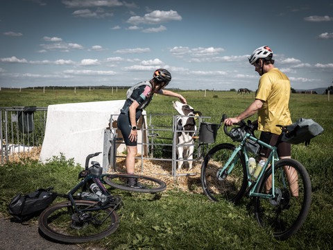 Dva cyklisté na šotolině RockHead na mléčné farmě Fiedler Zwei Radfahrer in Sportkleidung stehen auf einer Wiese; eine Person streichelt ein Kalb im Gatter, während die andere neben einem türkisfarbenen Fahrrad steht.Two cyclists in sportswear are standing in a meadow; one person is stroking a calf in a fence while the other is standing next to a turquoise-colored bicycle.Dva cyklisté ve sportovním oblečení stojí na louce; jeden hladí tele v ohradě, zatímco druhý stojí vedle tyrkysově zbarveného kola.Dwóch rowerzystów w strojach sportowych stoi na łące; jedna osoba głaszcze cielaka w płocie, podczas gdy druga stoi obok roweru w kolorze turkusowym.Twee fietsers in sportkleding staan in een weiland; de een aait een kalf in een omheining terwijl de ander naast een turquoise gekleurde fiets staat.Due ciclisti in abbigliamento sportivo sono in piedi in un prato; una persona accarezza un vitello in un recinto mentre l'altra è in piedi accanto a una bicicletta di colore turchese.