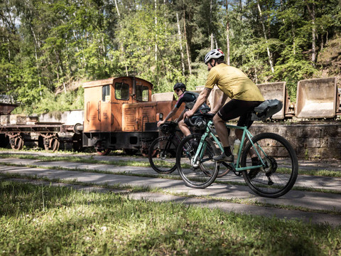 RockHead gravel bikers v muzeu lehké železnice Herrenleite Zwei Radfahrer mit Helmen fahren auf einem Weg neben einer alten, orangefarbenen Lokomotive, umgeben von grünen Bäumen.
