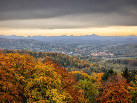 Ausblick vom Ungerberg im Herbst Herbstliche Landschaft mit bunten Bäumen im Vordergrund, einem Dorf in der Mitte und Bergen im Hintergrund unter bewölktem Himmel.