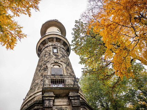Prinz-Georg-Turm auf dem Ungerberg Steinerner Turm mit Balkon, umgeben von herbstlichen Bäumen mit gelben Blättern, unter bewölktem Himmel.