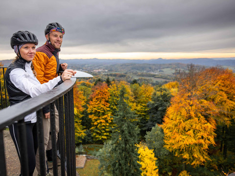 Zwei Gravelbiker auf dem Prinz-Georg-Turm/Ungerberg Zwei Personen mit Fahrradhelmen stehen an einem Geländer und blicken auf eine herbstliche Landschaft mit bunten Bäumen und bewölktem Himmel.