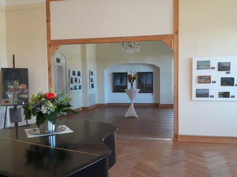 Innenbereich Schloss-Struppen Ein heller Ausstellungsraum mit Holzboden, Gemälden an den Wänden, einem Blumenarrangement auf einem Tisch und einem Kronleuchter an der Decke.A bright showroom with a wooden floor, paintings on the walls, a flower arrangement on a table and a chandelier on the ceiling.Světlý showroom s dřevěnou podlahou, obrazy na stěnách, květinovým aranžmá na stole a lustrem na stropě.Jasny showroom z drewnianą podłogą, obrazami na ścianach, kompozycją kwiatową na stole i żyrandolem na suficie.Een lichte showroom met een houten vloer, schilderijen aan de muren, een bloemstuk op tafel en een kroonluchter aan het plafond.Uno showroom luminoso con pavimento in legno, quadri alle pareti, una composizione floreale su un tavolo e un lampadario sul soffitto.