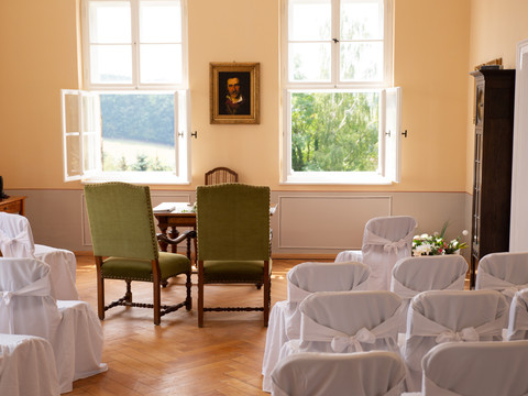 Hochzeitszimmer Schloss Struppen Heller Raum mit zwei grünen Stühlen vor einem Tisch, umgeben von weißen, mit Schleifen dekorierten Stühlen; Fenster mit Blick ins Grüne.Bright room with two green chairs in front of a table, surrounded by white chairs decorated with bows; window with a view of the greenery.Světlý pokoj se dvěma zelenými židlemi před stolem, obklopený bílými židlemi zdobenými mašlemi; okno s výhledem do zeleně.Jasny pokój z dwoma zielonymi krzesłami przed stołem, otoczony białymi krzesłami ozdobionymi kokardami; okno z widokiem na zieleń.Lichte kamer met twee groene stoelen voor een tafel, omringd door witte stoelen versierd met strikken; raam met uitzicht op het groen.Stanza luminosa con due sedie verdi davanti a un tavolo, circondato da sedie bianche decorate con fiocchi; finestra con vista sul verde.