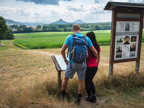 A couple stands in front of an information board on a hiking trail with a view of green fields and wooded hills under a cloudy sky.