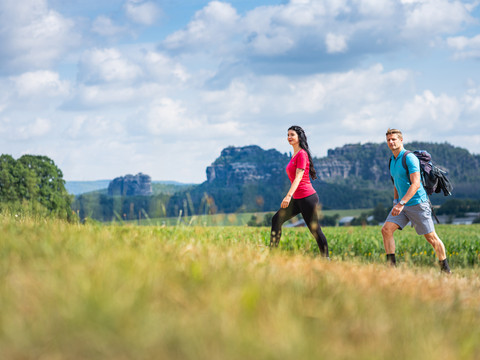 Two people are walking along a dirt track in front of a hilly landscape with rocks, the sky is cloudy. The woman is wearing a pink shirt, the man a blue one.