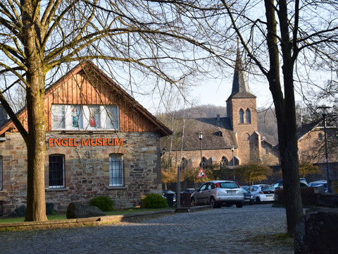 Engel-Museum Engel-Museum aus Backstein in Engelskirchen, mit Kirchturm im Hintergrund. Bäume rahmen die Szene ein.