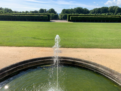 Parkanlage Barockgarten Großsedlitz Springbrunnen im Barockgarten Großsedlitz mit grüner Wiese und geschnittenen Hecken im Hintergrund unter blauem Himmel.