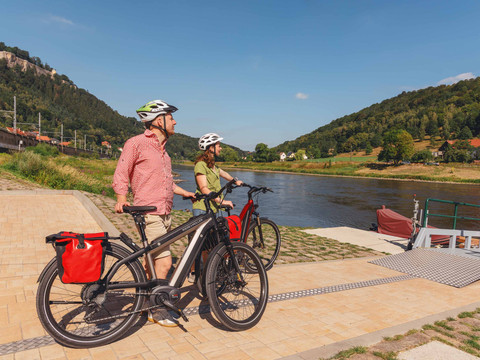 At the Königstein ferry pier Zwei Personen mit Helmen stehen neben E-Bikes mit roten Taschen an einem Flussufer, umgeben von grünen Hügeln und blauem Himmel.Two cyclists at the Königstein ferry pierDva cyklisté u přístaviště trajektu KönigsteinTwee fietsers bij de veerboothaven KönigsteinDwie osoby w kaskach stoją obok rowerów elektrycznych z czerwonymi torbami na brzegu rzeki, otoczone zielonymi wzgórzami i błękitnym niebem.Due persone con il casco sono in piedi accanto a biciclette elettriche con borse rosse sulla riva di un fiume, circondate da colline verdi e cielo blu.