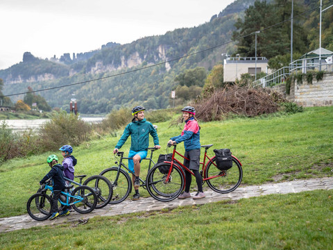 Familie met fietsen bij de Schmilka veerboot Eine Familie mit zwei Erwachsenen und zwei Kindern auf Fahrrädern, trägt Helme und bunte Kleidung, steht auf einem Weg in einer grünen, hügeligen Landschaft.Family with children and bikes waiting for the ferry in SchmilkaRodina s dětmi a koly čekající na trajekt ve SchmilceGezin met kinderen en fietsen wachtend op de veerboot in Schmilka