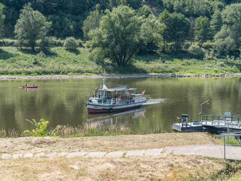 Ferry Schmilka Ein kleines Boot fährt auf einem ruhigen Fluss, während im Hintergrund ein Kanu mit zwei Personen paddelt; umgeben von grüner Landschaft.A small boat sails on a calm river, while a canoe with two people paddles in the background, surrounded by a green landscape.Malá loďka pluje po klidné řece, zatímco v pozadí pádluje kánoe se dvěma lidmi obklopená zelenou krajinou.Mała łódka płynie po spokojnej rzece, podczas gdy kajak z dwiema osobami wiosłuje w tle, otoczony zielonym krajobrazem.Een klein bootje vaart op een kalme rivier, terwijl een kano met twee personen peddelt op de achtergrond, omgeven door een groen landschap.Una piccola barca naviga su un fiume tranquillo, mentre una canoa con due persone pagaia sullo sfondo, circondata da un paesaggio verde.