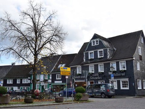 Ortskern Witzhelden Fachwerkhausplatz in Leichlingen mit Café, Bäckerei, herbstlichem Baum und parkendem Lieferwagen.