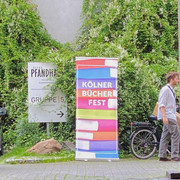 Cologne Book Festival Ein buntes Werbebanner für das Kölner Bücherfest steht vor einer bewachsenen Wand, daneben ein Fahrrad.A colorful advertising banner for the Cologne Book Festival stands in front of an overgrown wall, with a bicycle next to it.