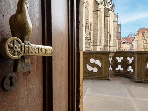 Rathaus Osnabrück Deur met decoratieve deurklink naast een stenen balkon en historische gebouwen op de achtergrond.