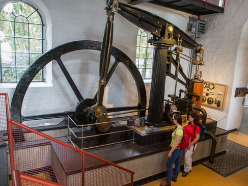 Zwei Personen betrachten eine historische Dampfmaschine mit Schwungrad in einem Industriemuseum.