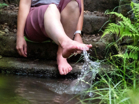 Waldlehrpfad mit Tretbecken Person in rosa Hose, sitzt auf Treppe, taucht Füße in sprudelndes Bachwasser, umgeben von Farnen.
