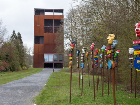 Rostiger Turm neben bunten, bemalten Maskenpfählen auf einem Weg durch Bäume.