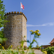 _BBB7566.jpeg Runder Burgturm aus Stein mit Zinnen und wehender rot-weißer Fahne bei klarem Himmel.
