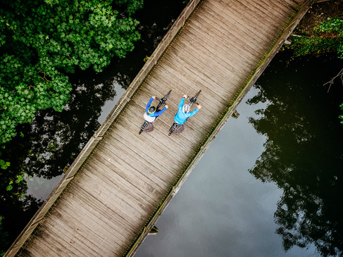 Zwei Radfahrer auf einer Holzbrücke über einem ruhigen Fluss, umgeben von grünen Bäumen.Two cyclists on a wooden bridge over a quiet river, surrounded by green trees.To cyklister på en træbro over en stille flod, omgivet af grønne træer.Twee fietsers op een houten brug over een rustige rivier, omringd door groene bomen.