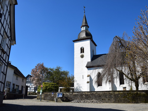 Bonte Kerke in Nümbrecht - Marienberghausen <p>Katholische Kirche mit weißer Fassade und Turm, umgeben von Fachwerkhäusern unter blauem Himmel.</p>
