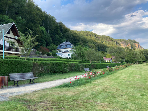 Ein gepflegter Park mit Bänken und Blumenbeeten, umgeben von grünen Hecken und Bäumen, im Hintergrund Häuser und ein bewaldeter Hügel.
