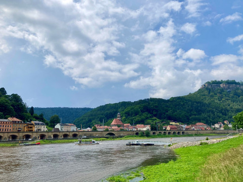 Trajekt Königstein_Halbestadt Flusslandschaft mit Brücke und bunten Häusern im Vordergrund, bewaldete Hügel im Hintergrund, teils bewölkter Himmel.
