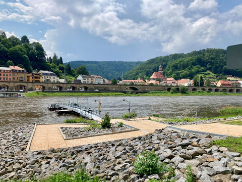 Veerboot Königstein_Halbestadt Toegang Flusslandschaft mit einer Steinbrücke und historischen Gebäuden im Hintergrund, umgeben von grünen Hügeln unter bewölktem Himmel.River landscape with a stone bridge and historic buildings in the background, surrounded by green hills under a cloudy sky.Říční krajina s kamenným mostem a historickými budovami v pozadí, obklopená zelenými kopci pod zataženou oblohou.Krajobraz rzeki z kamiennym mostem i zabytkowymi budynkami w tle, otoczony zielonymi wzgórzami pod zachmurzonym niebem.Rivierlandschap met een stenen brug en historische gebouwen op de achtergrond, omgeven door groene heuvels onder een bewolkte hemel.Paesaggio fluviale con ponte in pietra ed edifici storici sullo sfondo, circondato da colline verdi sotto un cielo nuvoloso.