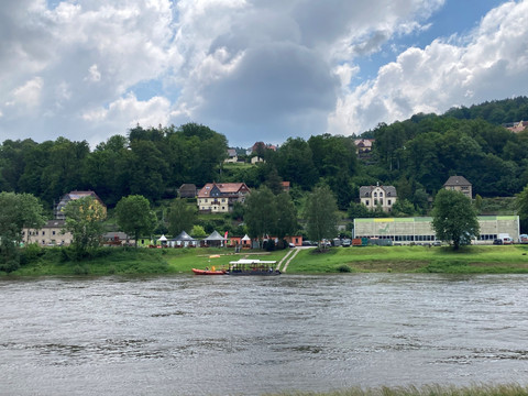 Kanu Aktiv Tours Flusslandschaft mit bewaldetem Hügel im Hintergrund, mehreren Häusern und einem Zelt. Der Himmel ist bewölkt, die Atmosphäre wirkt ruhig.