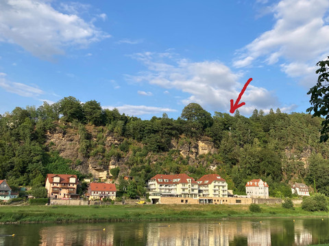 Klein bastion in Kurort Rathen Häuser mit roten Dächern am Fuße eines bewaldeten Hügels, darüber blauer Himmel mit Wolken; ein roter Pfeil zeigt auf einen Baum.
