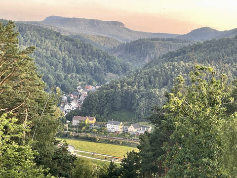Ausblick vom Skywalk  Blick auf ein Dorf in einem bewaldeten Tal, umgeben von grünen Hügeln und Bäumen, bei sanftem Abendlicht.