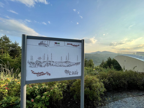 Erklärtafel  Ein Schild mit einer Karte der "Kuranlagen Ostrauer Ring" steht vor einer grünen Landschaft und einem modernen Gebäude unter blauem Himmel.
