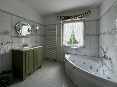 Haus Thal - Zimmer 2 - Badezimmer Das Bild zeigt ein helles Badezimmer mit einer Eckbadewanne, Waschbecken und Fenster mit Vorhängen.