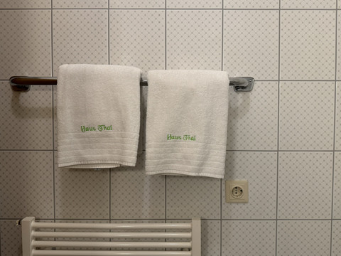 Haus Thal - Handtücher mit Logo Zwei weiße Handtücher auf einem Halter vor einer gefliesten Wand im Badezimmer, eine Steckdose sichtbar.