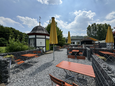Haus Thal - Ansicht Außenbereich Ein Biergarten mit orangefarbenen Tischen und gelben Sonnenschirmen auf Kiesboden, umgeben von Grünflächen.
