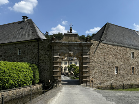 Schloss Ehreshoven  Ein steinernes Tor führt zu einem historischen Anwesen, umgeben von Bäumen und sonnigem Himmel.