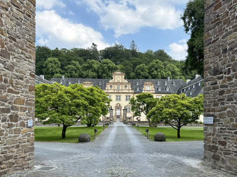 Schloss Ehreshoven Barockes Herrenhaus mit gelber Fassade, flankiert von Bäumen, hinter einem Torbogen, im Sommer.
