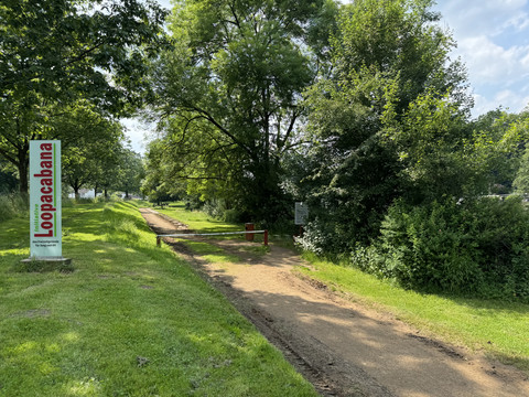 Loopacabana Ruhiger, grüner Parkweg gesäumt von Bäumen und Büschen, mit Loopacubana-Schild am Wegrand.