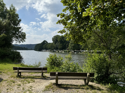Freizeitanlage Loopacabana Zwei Holzbänke am ruhigen Seeufer, umgeben von Bäumen, mit Blick auf das grüne Ufer gegenüber.