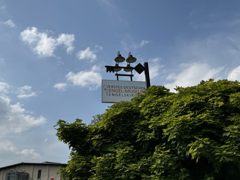 Engel-Museum Ein Schild des Ersten Deutschen Engel-Museums in Engelskirchen, umgeben von grünem Laub vor blauem Himmel.