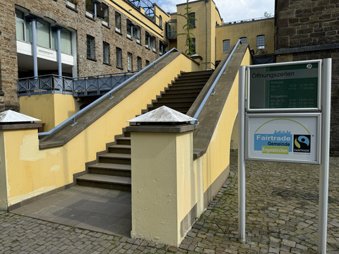 LVR-Industriemuseum Stufenaufgang zu einem gelben Gebäudekomplex, daneben ein Schild mit Öffnungszeiten und Fairtrade-Hinweis.