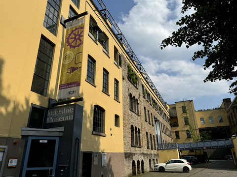 LVR-Industriemuseum Gelbes Gebäude eines Industriemuseums mit blauen Fenstern, Werbeschildern und einem weißen Auto davor.