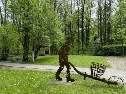 Papiermuseum Metallskulptur eines Mannes mit Schubkarre, umgeben von grünen Bäumen und Rasen im Park.