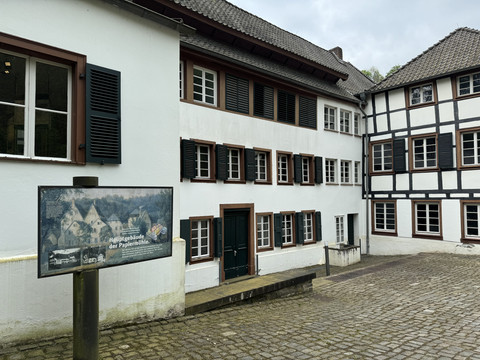 Papiermuseum Alte Dombach Fachwerkhäuser mit Holzläden, gepflasterter Hof und Informationstafel bei bewölktem Himmel.