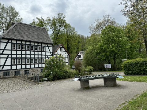 Papiermuseum  Fachwerkhäuser inmitten grüner Bäume und ein modernes Kunstwerk auf einer gepflasterten Fläche.