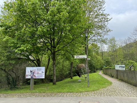 Papiermuseum Grüne Parkanlage mit Info-Tafel, Kunstskulptur und umgebenden Bäumen an einem Gehweg.