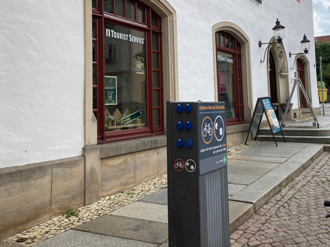 Pirna e-bike oplaadpunt.jpg Ladestation für E-Bikes vor einem Gebäude mit der Aufschrift "Tourist Service", rote Fensterrahmen, zwei Laternen und Infotafeln auf dem Gehweg.