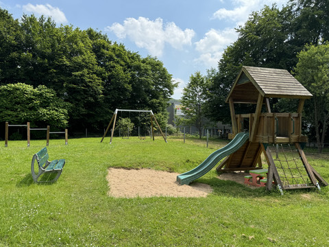 Haus Thal - Ansicht Spielplatz Spielplatz mit Kletterturm, Rutsche, Schaukel und Holzsitzbank, umgeben von üppigem Grün.