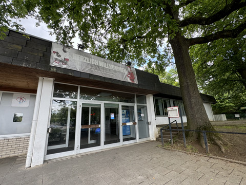 Eissportarena Eingangsbereich eines Gebäudes mit großen Glasfenstern, Willkommensschild und einem großen Baum daneben.